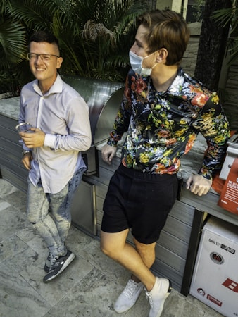 Two men are standing near a grill in a garden area. One man is smiling, wearing glasses, a light-colored shirt, and denim jeans while holding a drink. The other man is wearing a floral shirt, black shorts, and a mask, leaning casually. Lush green foliage surrounds them.