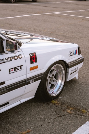 The image captures a close-up view of a white sports car parked on an asphalt surface. The car features various decals and logos such as Nissan PDC, FET Kyokuto, SSR, and HKS. The focus is on the rear section of the car, showcasing the distinctive rims and the sleek bodywork.