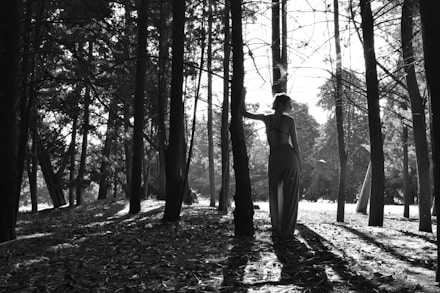 A woman holding a shamanic drum in a forest clearing with sunlight filtering through the trees.