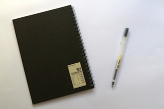 A black spiral-bound notebook with a label in the bottom right corner is placed next to a transparent pen with black ink. Both items are on a plain white surface.