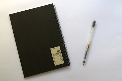 A black spiral-bound notebook with a label in the bottom right corner is placed next to a transparent pen with black ink. Both items are on a plain white surface.
