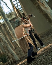 Two individuals in a wooded area engage in a dynamic action scene. One person, clad in a fur-covered lower garment with animal ears, is armed with two swords. The other person, wearing dark pants and a stained vest, wields large metal claws. The background consists of tree trunks, and the ground is covered with leaves.