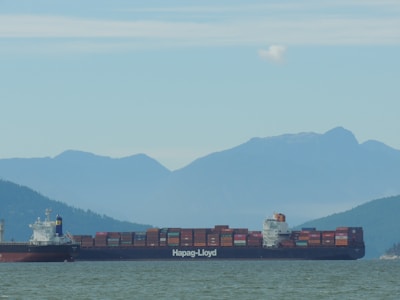 A large container ship sails through calm waters with a mountainous background. The ship is loaded with numerous multicolored shipping containers and bears the name Hapag-Lloyd. The misty mountains and cloudy sky create a serene and expansive atmosphere.