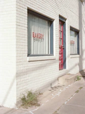 A brick building with large windows that read 'BAKERY' and 'BISTRO' in red lettering. The building has a white, painted facade with a red door. There are some green plants growing at the base of the building on the sidewalk, which also has some chalk or spray-painted markings.