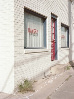A brick building with large windows that read 'BAKERY' and 'BISTRO' in red lettering. The building has a white, painted facade with a red door. There are some green plants growing at the base of the building on the sidewalk, which also has some chalk or spray-painted markings.