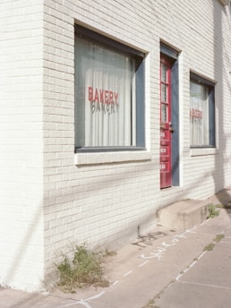 A brick building with large windows that read 'BAKERY' and 'BISTRO' in red lettering. The building has a white, painted facade with a red door. There are some green plants growing at the base of the building on the sidewalk, which also has some chalk or spray-painted markings.