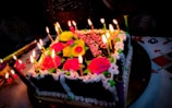 A square chocolate cake decorated with vibrant pink and yellow roses, surrounded by lit candles. The cake has white icing borders, and it is placed on a decorated tablecloth. Additional decorations include various colored candles and a festive atmosphere.