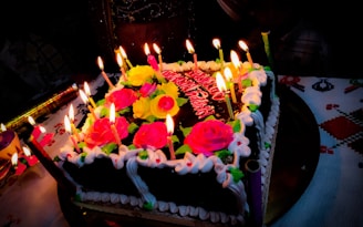 A square chocolate cake decorated with vibrant pink and yellow roses, surrounded by lit candles. The cake has white icing borders, and it is placed on a decorated tablecloth. Additional decorations include various colored candles and a festive atmosphere.