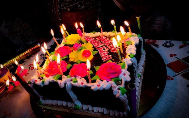 A square chocolate cake decorated with vibrant pink and yellow roses, surrounded by lit candles. The cake has white icing borders, and it is placed on a decorated tablecloth. Additional decorations include various colored candles and a festive atmosphere.