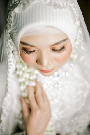 A close-up view of a person wearing a white hijab adorned with intricate lace and applique details. The face is softly illuminated, emphasizing long eyelashes and subtle makeup with warm tones. Hands are gently clasped, holding a delicate garland with white and green flowers. The expression appears serene and contemplative.