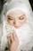 A close-up view of a person wearing a white hijab adorned with intricate lace and applique details. The face is softly illuminated, emphasizing long eyelashes and subtle makeup with warm tones. Hands are gently clasped, holding a delicate garland with white and green flowers. The expression appears serene and contemplative.