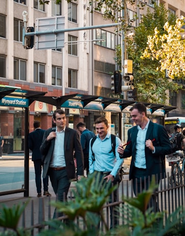 A busy urban scene with several people walking on a sidewalk near a bus stop labeled Wynyard Station and Stand H. Three individuals in business attire, holding bags, appear to be engaged in conversation. The backdrop includes tall office buildings and trees, suggesting a commercial area during daytime.