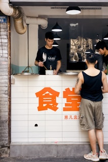 A small food stall is visible with a counter at the front, staffed by two individuals. One person is actively cooking or preparing food, while another stands nearby, possibly taking an order or waiting. A customer in a black tank top and khaki shorts stands in front of the counter. Large red Chinese characters are prominently displayed on the stall's frontage. Industrial-style hanging lights and ventilation pipes are noticeable in the background.