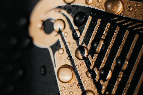 A close-up view of water droplets resting on a golden surface with black, textured lines running through it, suggesting a design or logo.