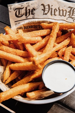 A plate of golden-brown French fries served on a piece of newspaper with a cup of creamy white dipping sauce on the side. The lighting highlights the crispy texture of the fries.