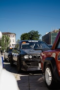 A lineup of modified racing cars parked on a city street, with their vibrant colors and numbers displayed on the windshields. The cars stretch back into the distance under a clear blue sky, surrounded by urban buildings and greenery.