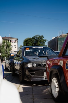 A lineup of modified racing cars parked on a city street, with their vibrant colors and numbers displayed on the windshields. The cars stretch back into the distance under a clear blue sky, surrounded by urban buildings and greenery.
