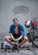 A man is sitting on a small stool, sharpening a blade on a stone. He is wearing a blue shirt open at the front, with shorts, and appears focused on his task. A bicycle is parked beside him, carrying a bag loaded with items. The setting is a tiled area, and various tools and objects are scattered on the ground nearby.