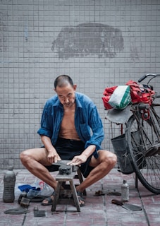 A man is sitting on a small stool, sharpening a blade on a stone. He is wearing a blue shirt open at the front, with shorts, and appears focused on his task. A bicycle is parked beside him, carrying a bag loaded with items. The setting is a tiled area, and various tools and objects are scattered on the ground nearby.