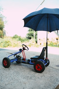 A go-kart with a blue frame and red wheels is parked on a concrete surface. An umbrella is attached to the kart, providing shade. The scene is outdoors with some greenery and a clear sky in the background.
