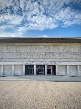A large, imposing building with classical architecture, featuring a wide set of steps leading up to the entrance. It has tall columns before the entryway and intricate carvings above depicting historical scenes. The facade has inscriptions in multiple languages.