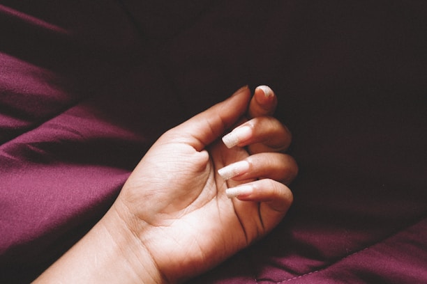 Close-up of a hand with freshly manicured nails in soft mauve tones, resting on a violet fabric.