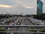 A busy urban scene with heavy traffic on multiple lanes of a broad highway. Numerous cars, buses, and auto-rickshaws are visible, along with a few motorbikes. The vehicles are predominantly stuck in a traffic jam on one side of the road, while the opposite lane shows relatively free movement. A tall, modern glass building stands prominently on the right side, while other urban structures and greenery are visible around the road. The sky is overcast with a hint of sunlight peeking through the clouds.