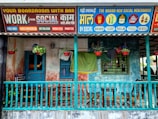 A colorful storefront featuring a wooden porch with a teal railing, furniture like chairs and a small table, and hanging plants in red pots. Above the porch, vibrant signs advertise various services and merchandise under the brand 'Social'. The signs are in a mix of English and another language, offering amenities like a boardroom with a bar, free WiFi, video conferencing, and merchandise such as beer glasses and pickle jars.