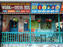 A colorful storefront featuring a wooden porch with a teal railing, furniture like chairs and a small table, and hanging plants in red pots. Above the porch, vibrant signs advertise various services and merchandise under the brand 'Social'. The signs are in a mix of English and another language, offering amenities like a boardroom with a bar, free WiFi, video conferencing, and merchandise such as beer glasses and pickle jars.