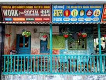 A colorful storefront featuring a wooden porch with a teal railing, furniture like chairs and a small table, and hanging plants in red pots. Above the porch, vibrant signs advertise various services and merchandise under the brand 'Social'. The signs are in a mix of English and another language, offering amenities like a boardroom with a bar, free WiFi, video conferencing, and merchandise such as beer glasses and pickle jars.