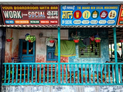A colorful storefront featuring a wooden porch with a teal railing, furniture like chairs and a small table, and hanging plants in red pots. Above the porch, vibrant signs advertise various services and merchandise under the brand 'Social'. The signs are in a mix of English and another language, offering amenities like a boardroom with a bar, free WiFi, video conferencing, and merchandise such as beer glasses and pickle jars.