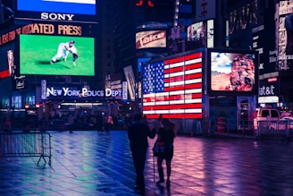 A vibrant city scene featuring numerous bright electronic billboards and signage, including a large American flag display and an advertisement for the New York Police Department. The ground glistens with reflections from the lights, and two people are walking through the square.