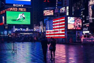 A vibrant city scene featuring numerous bright electronic billboards and signage, including a large American flag display and an advertisement for the New York Police Department. The ground glistens with reflections from the lights, and two people are walking through the square.