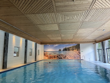 A spacious indoor swimming pool with clear blue water is set against a backdrop of white tiled walls. The wooden ceiling has a geometric pattern. Large windows on one side let in natural light, and a mural of a serene beach scene adds an element of relaxation.