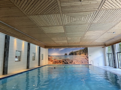 A spacious indoor swimming pool with clear blue water is set against a backdrop of white tiled walls. The wooden ceiling has a geometric pattern. Large windows on one side let in natural light, and a mural of a serene beach scene adds an element of relaxation.