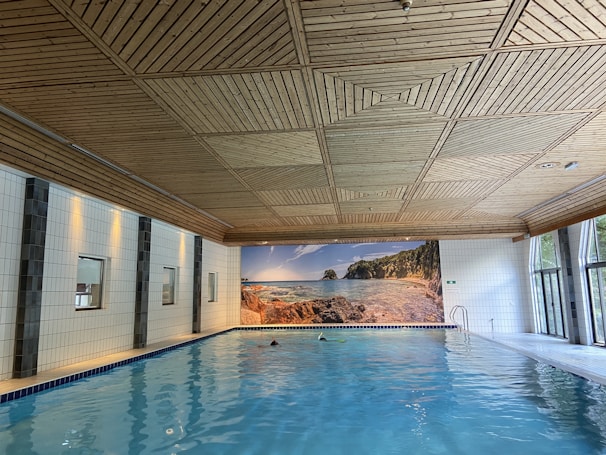 A spacious indoor swimming pool with clear blue water is set against a backdrop of white tiled walls. The wooden ceiling has a geometric pattern. Large windows on one side let in natural light, and a mural of a serene beach scene adds an element of relaxation.