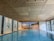 A spacious indoor swimming pool with clear blue water is set against a backdrop of white tiled walls. The wooden ceiling has a geometric pattern. Large windows on one side let in natural light, and a mural of a serene beach scene adds an element of relaxation.