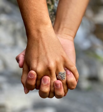 Two hands are holding each other, with one hand wearing an ornate ring featuring an intricate design. The background is softly blurred, focusing attention on the hands and the symbol of connection and affection they represent.