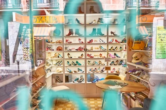 A shoe store display with shelves filled with a variety of colorful shoes, including heels and boots. The interior is visible through the store window with reflections, and there are hats and other items on side shelves. A small table and chairs are situated in the foreground, along with some crafting materials.