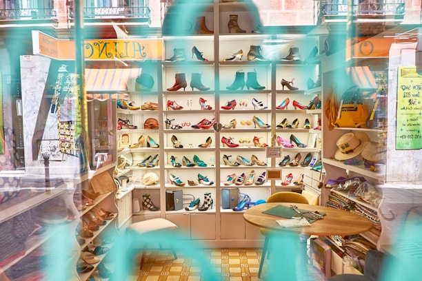 A shoe store display with shelves filled with a variety of colorful shoes, including heels and boots. The interior is visible through the store window with reflections, and there are hats and other items on side shelves. A small table and chairs are situated in the foreground, along with some crafting materials.