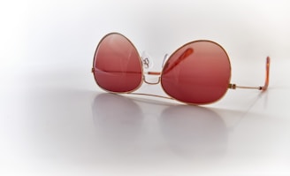 A pair of oversized sunglasses with large, round pink-tinted lenses and thin gold frames, resting on a reflective white surface. The lenses are semi-transparent, and the reflection is visible beneath the glasses.