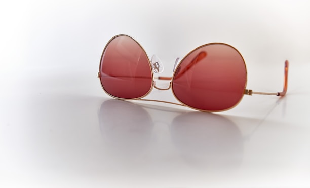 A pair of oversized sunglasses with large, round pink-tinted lenses and thin gold frames, resting on a reflective white surface. The lenses are semi-transparent, and the reflection is visible beneath the glasses.