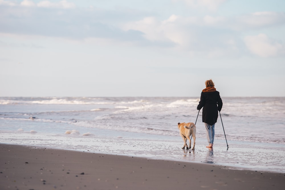 woman in black jacket walking with dog on beach during daytime