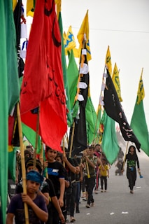 A group of people, mostly young, are participating in a procession, carrying vibrant flags in a variety of colors including red, green, yellow, and black. The flags have different symbols and inscriptions. Participants are walking closely together on a road under an overcast sky, conveying a sense of unity and movement.
