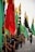 A group of people, mostly young, are participating in a procession, carrying vibrant flags in a variety of colors including red, green, yellow, and black. The flags have different symbols and inscriptions. Participants are walking closely together on a road under an overcast sky, conveying a sense of unity and movement.
