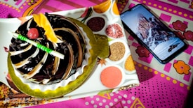 A chocolate cake with decorative swirls, garnished with a cherry and a piece of yellow fruit, sits on a decorative plate adorned with images of spices. Next to the cake, a smartphone displays an image of a snowy mountain landscape.