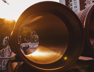 Large industrial pipes are stacked horizontally in an urban setting during sunset. The sunlight casts a warm glow through the pipes, creating a striking contrast with the surrounding architecture and trees.