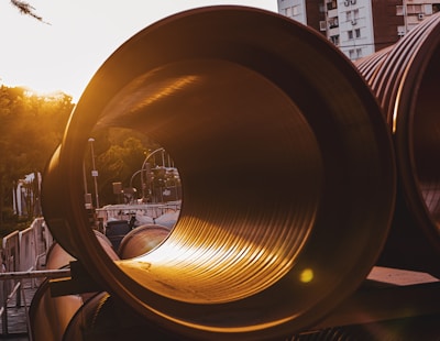 Large industrial pipes are stacked horizontally in an urban setting during sunset. The sunlight casts a warm glow through the pipes, creating a striking contrast with the surrounding architecture and trees.