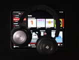 A pair of black over-ear headphones rests on top of a tablet displaying a music streaming app. Various music categories and album art are visible on the screen.