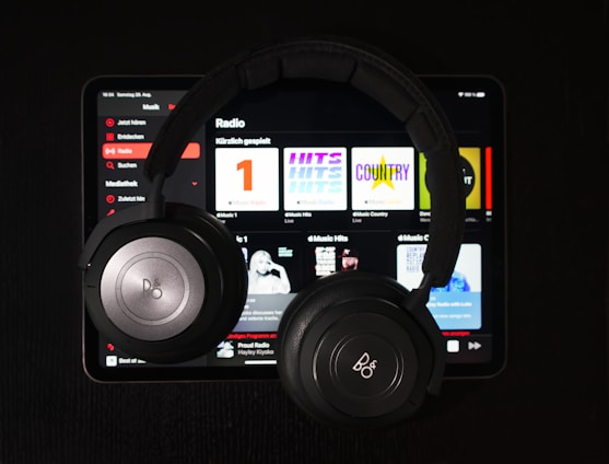 A pair of black over-ear headphones rests on top of a tablet displaying a music streaming app. Various music categories and album art are visible on the screen.